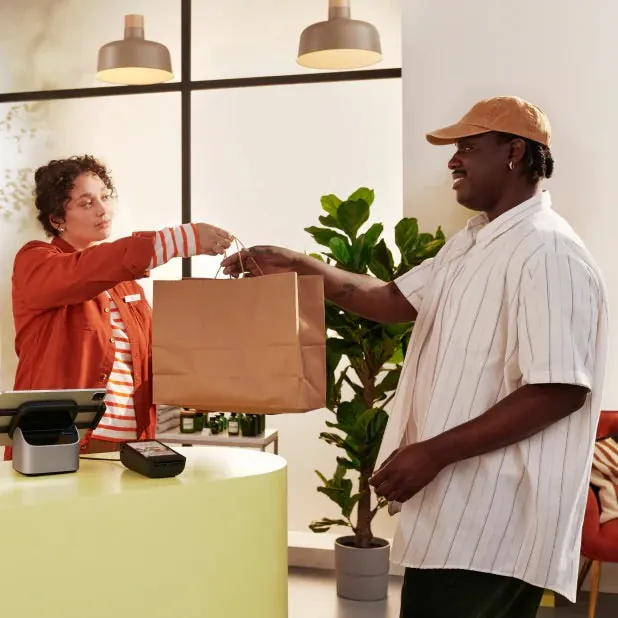 Shop assistant handing a paper bag to a customer across a counter with a POS terminal