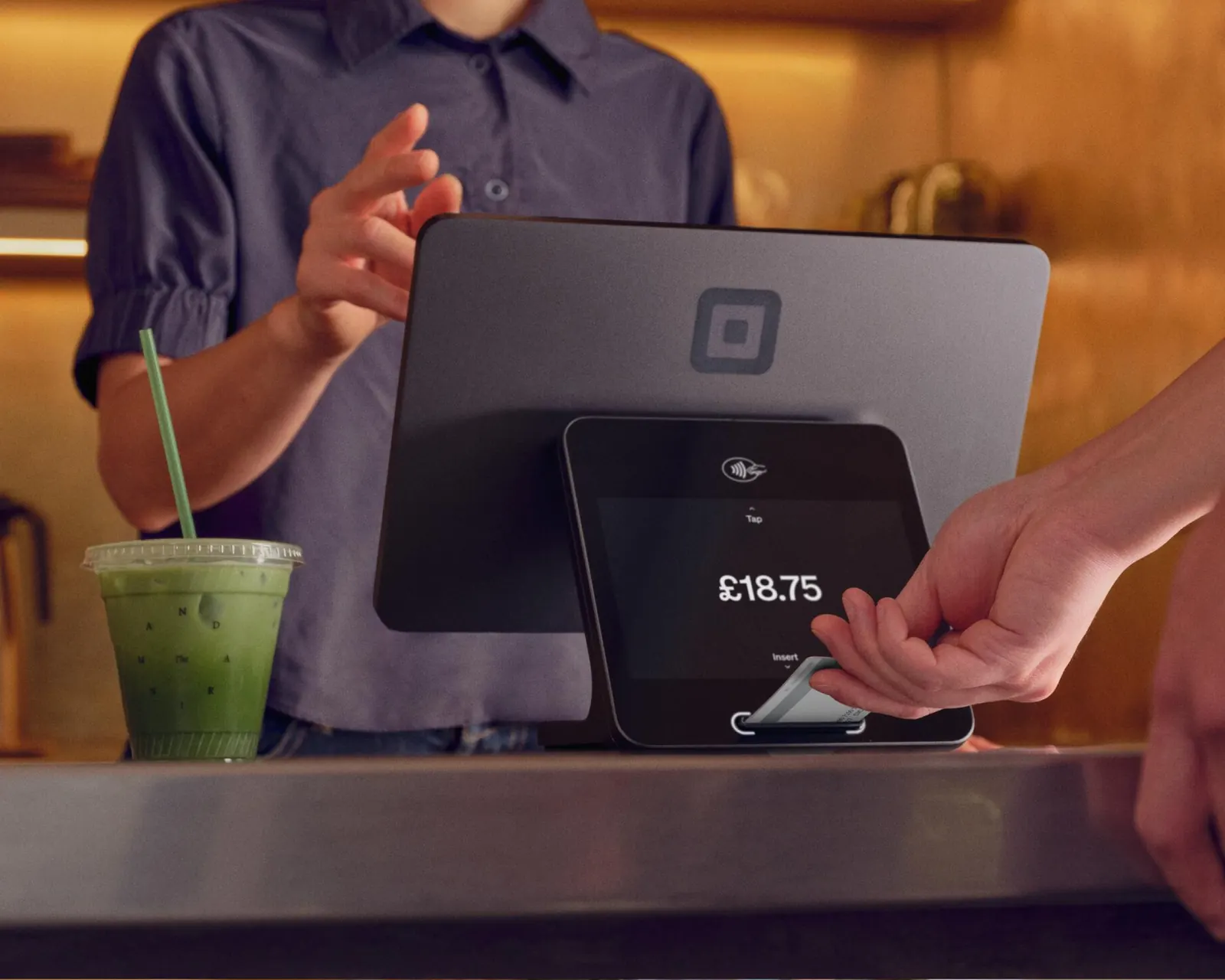 Cashier hand holding out a card to a Square Stand showing a payment total, next to a green drink