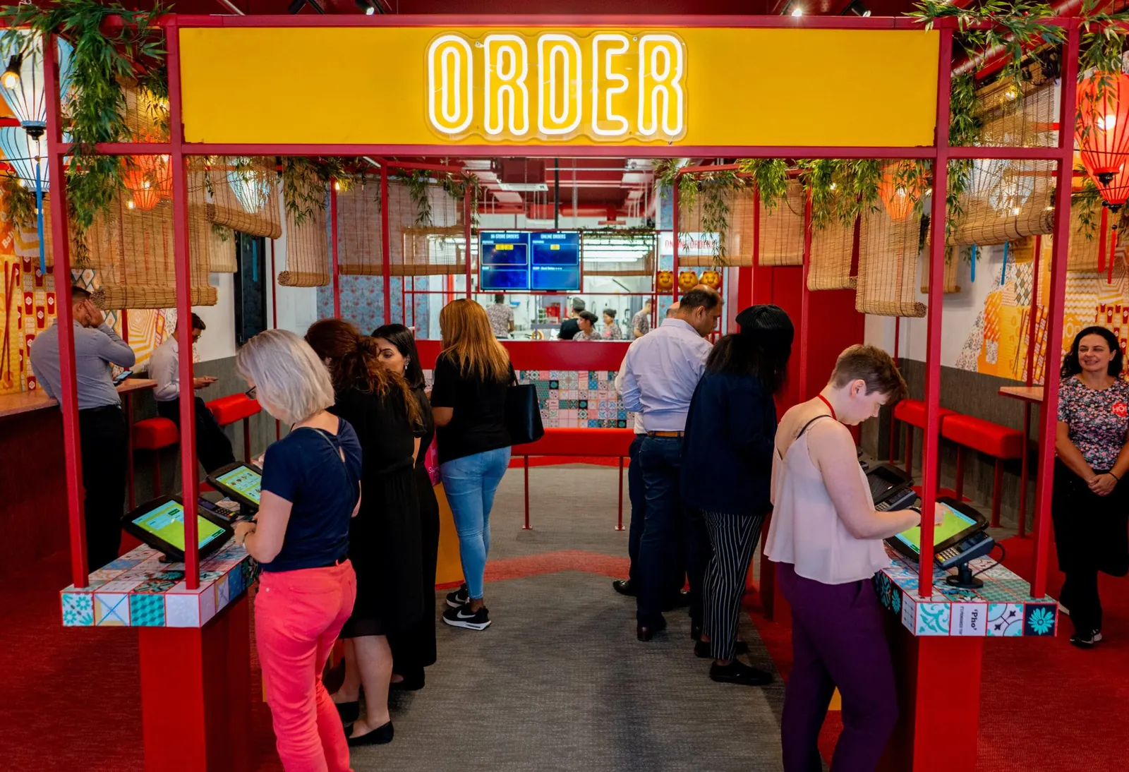 Customers queueing at a row of self-service kiosks under a neon ORDER sign