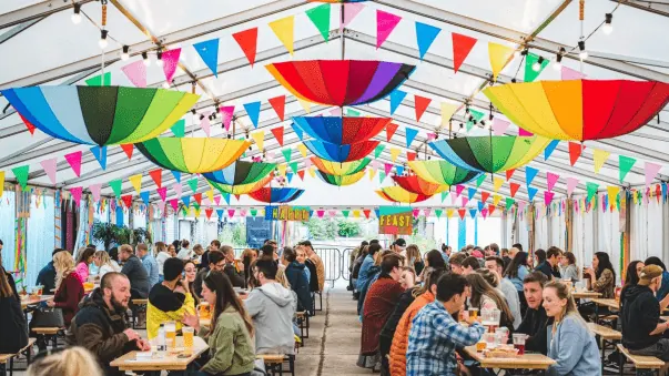 Crowd seated at long tables under a marquee hung with bunting and umbrellas